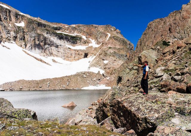 Volunteer Kevin Zagorda points to a glacier in Rocky Mountain National Park. (Kevin Zagorda) Man standing on rock beside mountain lake pointing to snowfield.