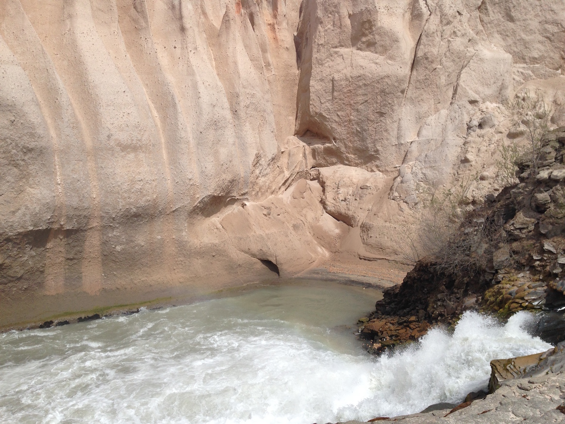 A low waterfall gushes into a pool below tall tan cliffs