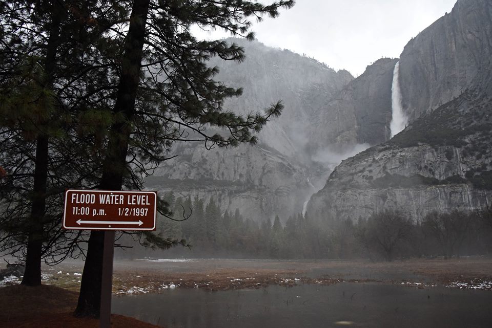 A historic flood marker sign in front of a flooded meadow and waterfall in the background.