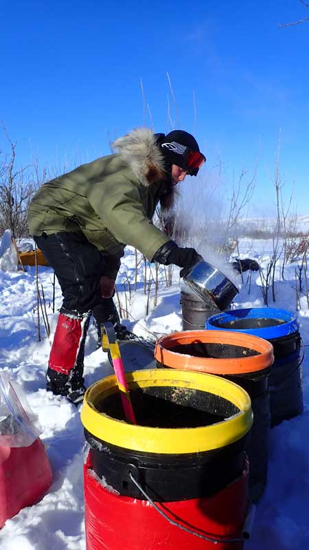 a woman in winter gear fills buckets with hot water and dog food