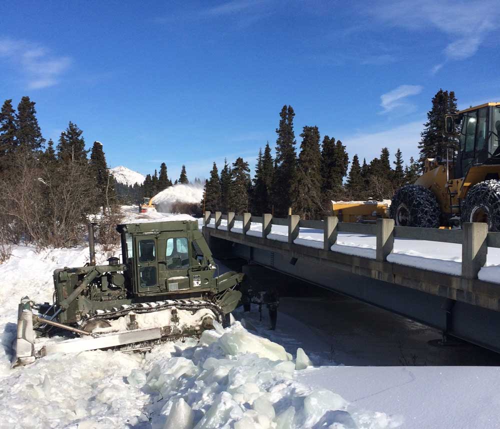 a green bulldozer ripping up sheets of ice below a bridge