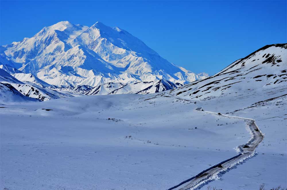 a snowy landscape with a plowed road leading in the direction of a vast snowy mountain