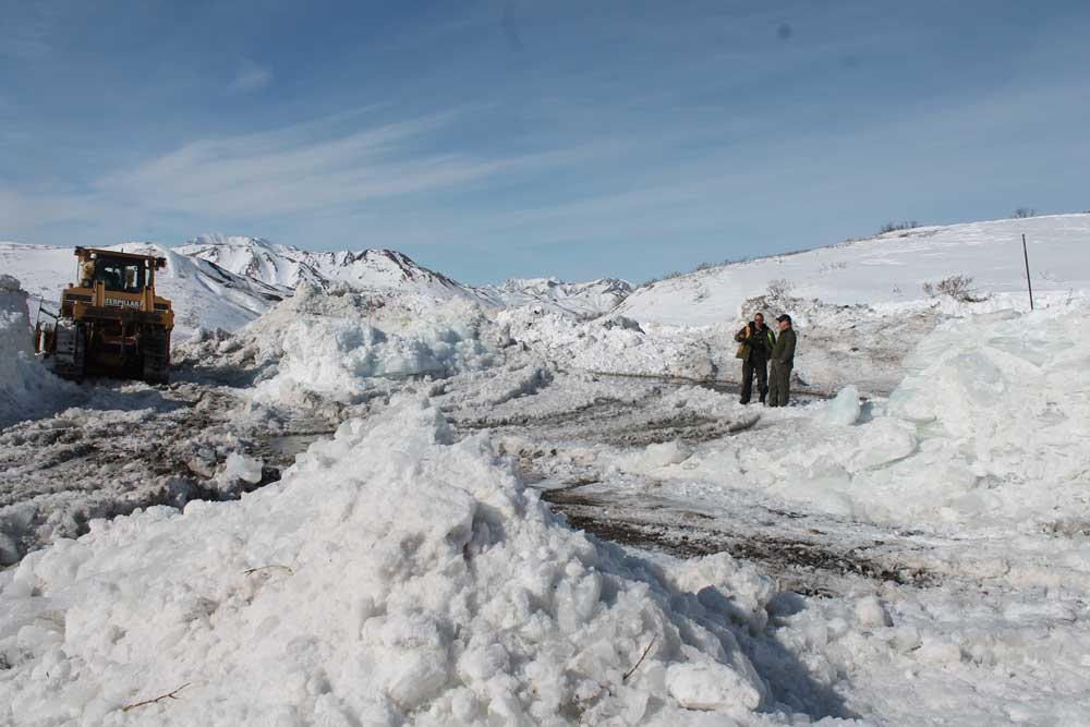 Mile 40.5 two men stand on a road surrounded by large piles of plowed snow, near a piece of heavy machinery