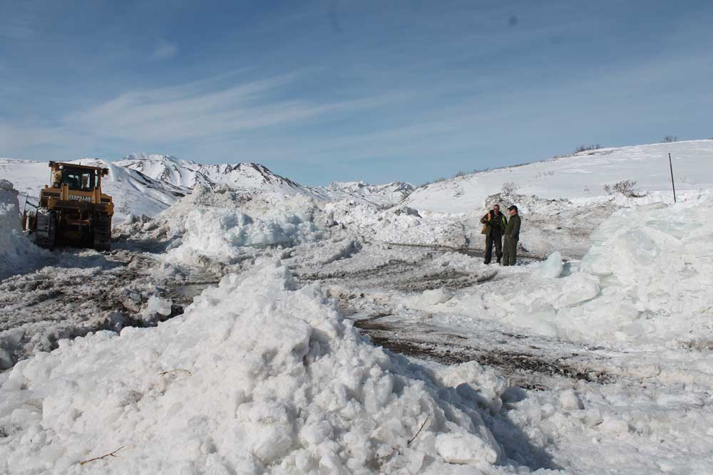 two men stand on a road surrounded by large piles of plowed snow, near a piece of heavy machinery