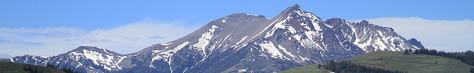 Photo of green rolling hills in the foreground with lingering snow on Electric Peak in the distance.