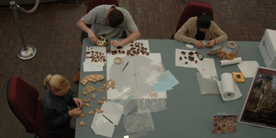 Color photo of three people seated at a table piecing together bits of pottery. Color photo of three people seated at a table piecing together bits of pottery.