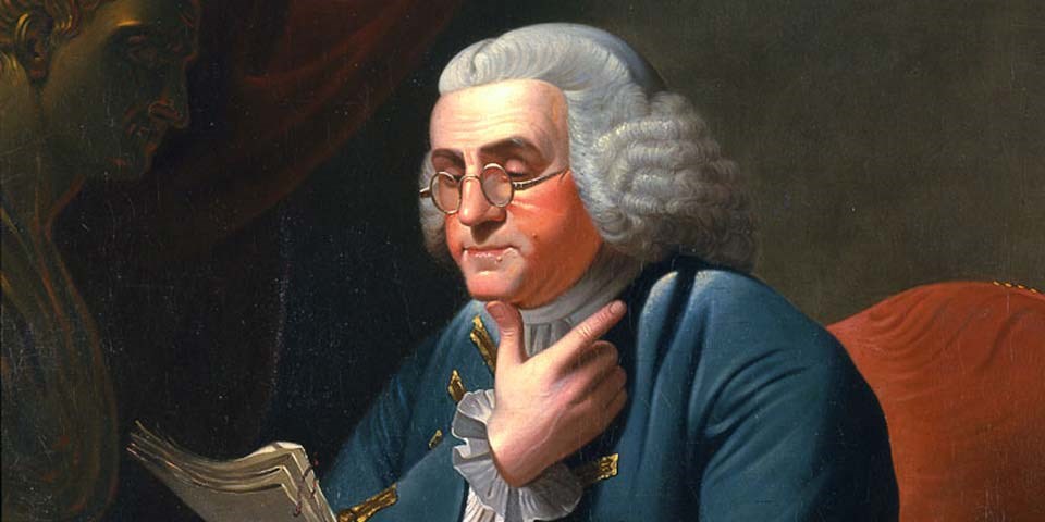 Color image of a detail of a painting of Benjamin Franklin, showing Franklin seated, wearing a powdered wig and silver rimmed glasses, with his chin resting on his right thumb.