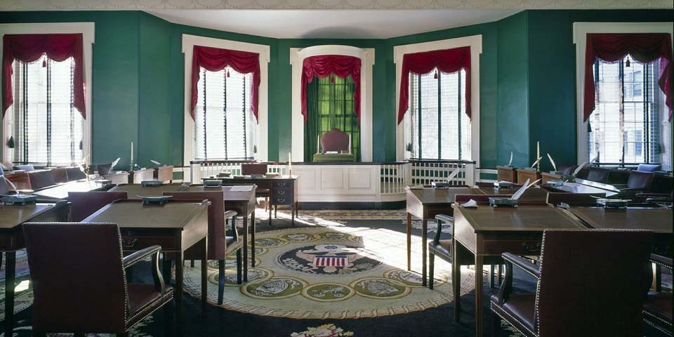 Color photo of the Senate chamber, with two rows of wooden desks facing a single chair on a raised dais that sits in front of large windows.