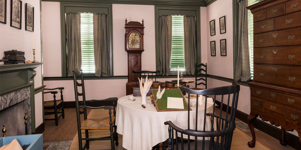 Color photo of the Declaration House parlor with a Windsor chair and table, tall chest, and tall case clock. Color photo of the Declaration House parlor with a Windsor chair and table, tall chest, and tall case clock.