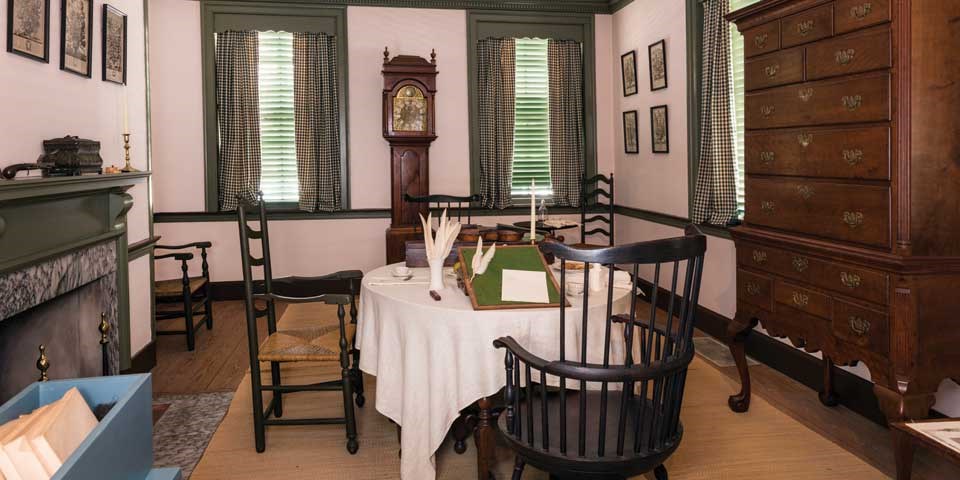 Color photo showing parlor with Windsor chairs around table in foreground, and tall case clock on pier wall in background. Color photo showing parlor with Windsor chairs around table in foreground, and tall case clock on pier wall in background.