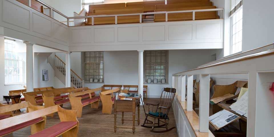 Color photo showing wooden pews facing a rocking chair in the front of the room with a balcony above.