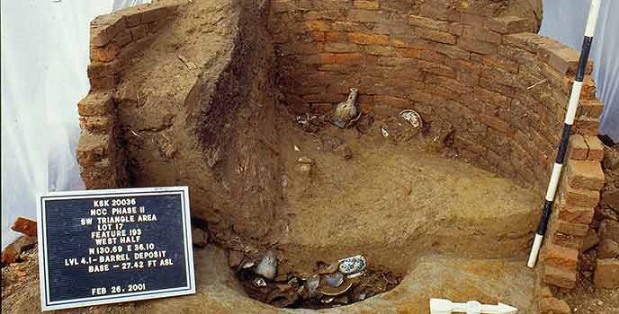 The photograph shows a partially excavated brick-lined pit, with artifacts that have settled to the bottom.