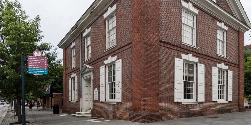 Color photo of the Free Quaker Meeting House exterior, a red brick building with white shutters situated on a corner lot.