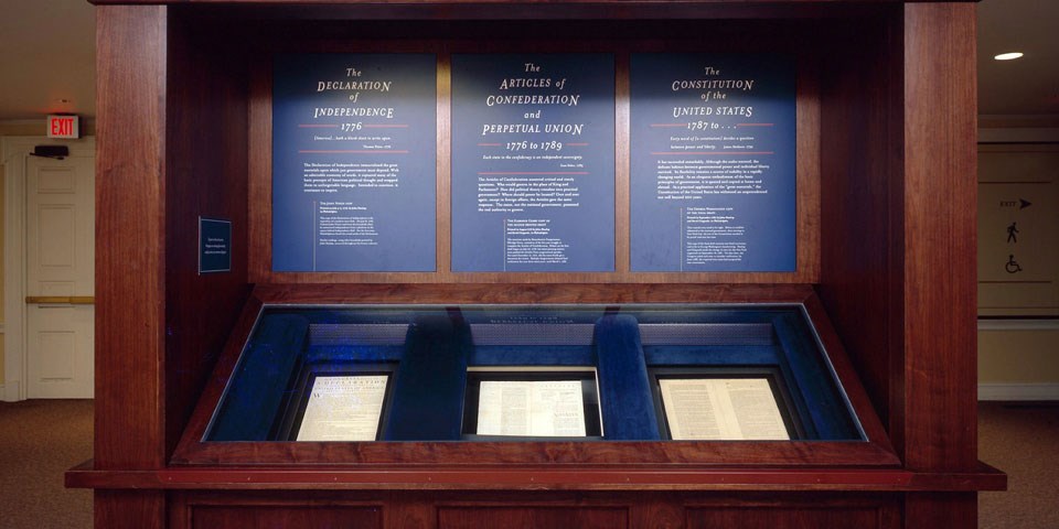 Color photo of a wooden exhibit case displaying three documents, side by side, at waist height with blue text panels above.