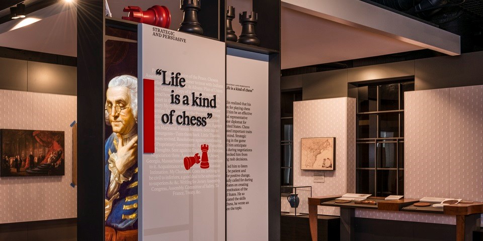 Color photo of an exhibit showing a reproduction painting of Benjamin Franklin next to a quote with red letters that states life is a kind of chess.