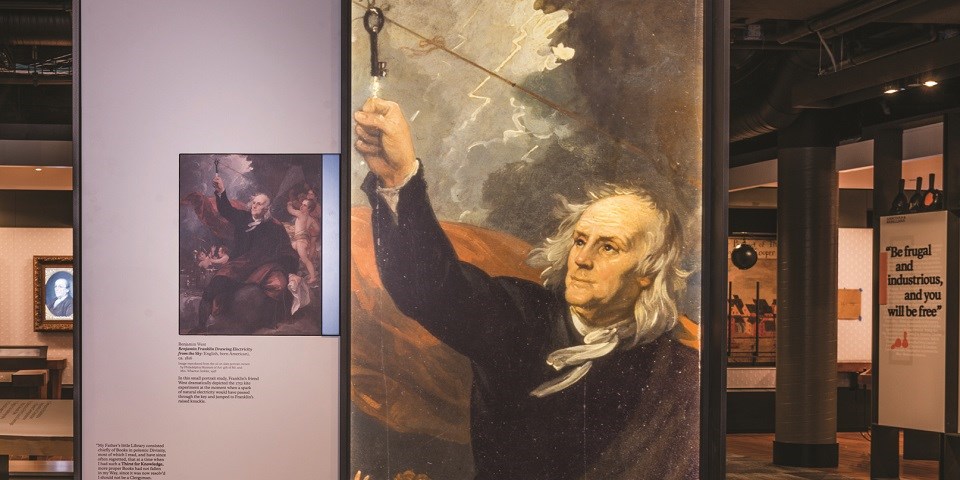 Color image of an exhibit with an image of an older Benjamin Franklin holding up his knuckle to touch a black key on a string.