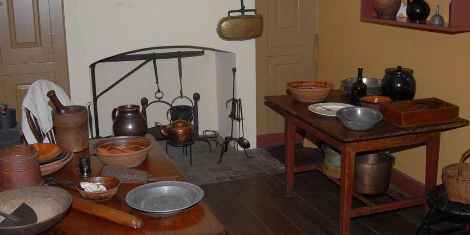 Color photo of the Todd House kitchen showing a narrow work space with wooden work tables on either side of the room and a fireplace along the back wall.  Redware bowls and pewter utensils sit on the tables and near the fireplace.
