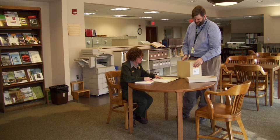 Color photo of an archivist putting a box of archival material on a table near a seated female park ranger. Color photo of an archivist putting a box of archival material on a table near a seated female park ranger.