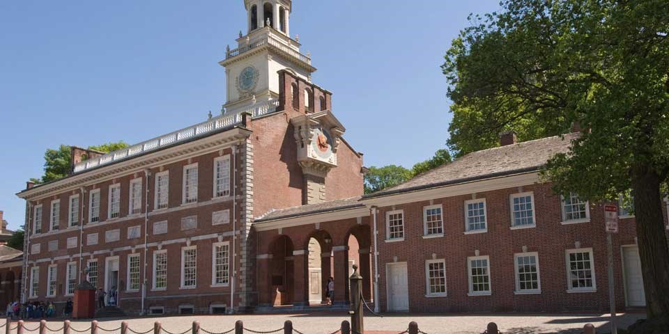 Color photo of Independence Hall and its West Wing, showing a large two story red brick building with central clock tower and adjoining smaller two story red brick building.