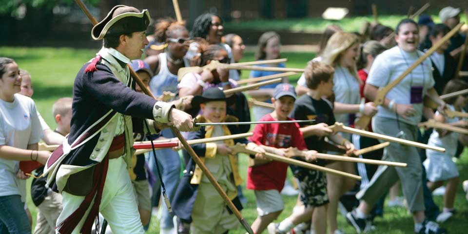 Color photo showing people of all ages running with wooden muskets while a man in colonial military uniform encourages them on.