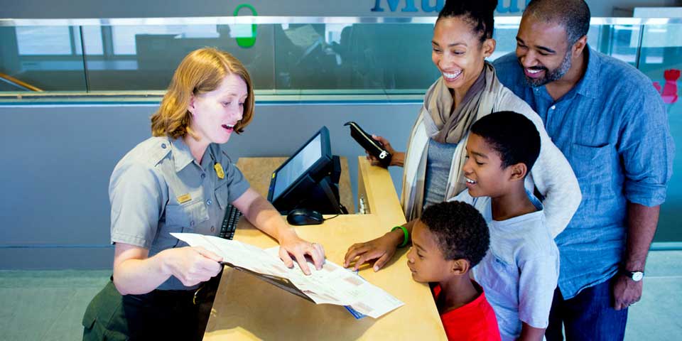 Color photo of a female park ranger at a desk holding up a map for a family to see.