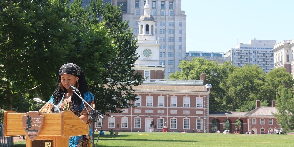 Color image of a young woman speaking at a podium with Independence Hall in the background.