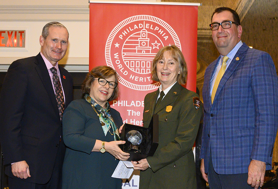 Globy Superintendent MacLeod is presented with the Globy award. There are two people on her right, one on her left. They are standing in front of a Global Philadelphia sign