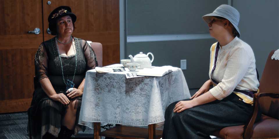 Color photo of two women wearing early 1900s attire having conversation at a tea table.