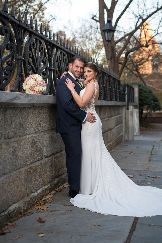 A picture of a couple in wedding clothes against a marble wall and iron fence.