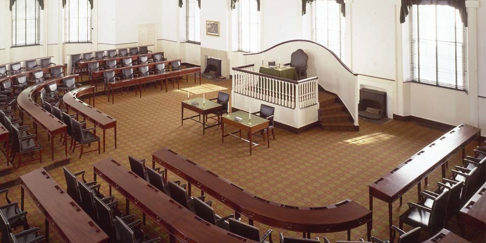 Color photo of the interior of Congress Hall, looking down on the House of Representatives with three rows of desks and chairs in a horseshoe shape facing a raised dais with one large chair.