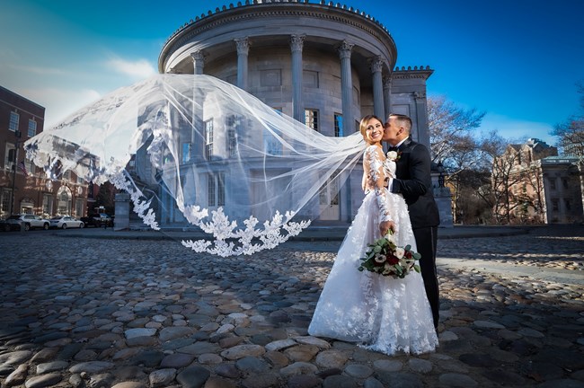 A couple in wedding clothes on a "cobblestone" street with a marble building and rotunda in background. The brides veil blows outward in wind.