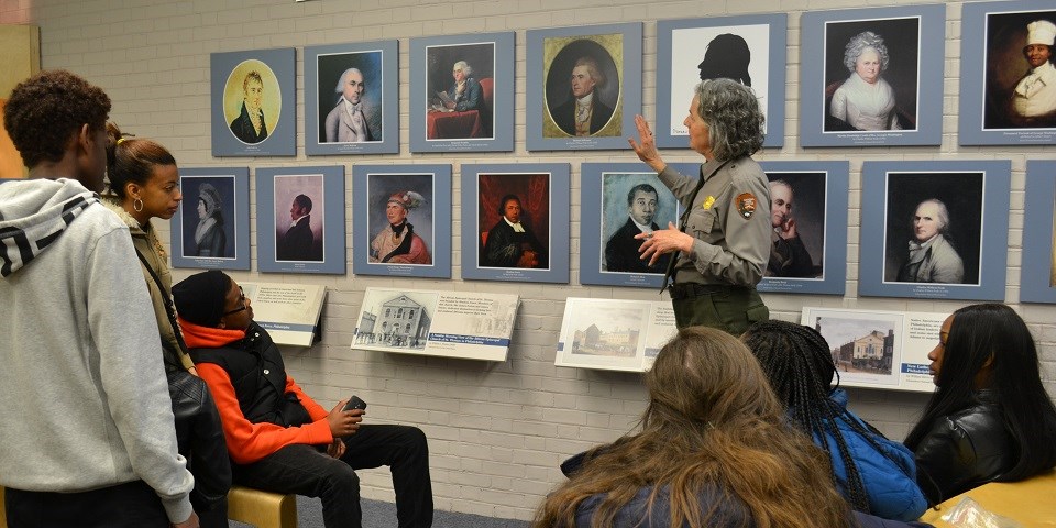 Color image showing a group of students looking at a wall with mounted 18th-century portraits while a female ranger speaks.