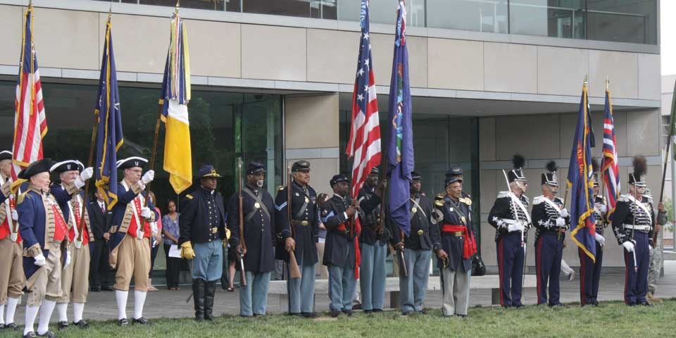 Color photo of reenactors dressed in military uniforms from different periods in history holding flags and standing in a line. Color photo of reenactors dressed in military uniforms from different periods in history holding flags and standing in a line.