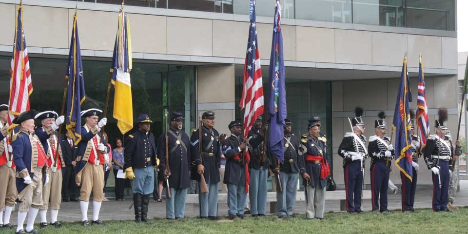 Color photo of reenactors dressed in military uniforms from different periods in history holding flags and standing in a line.