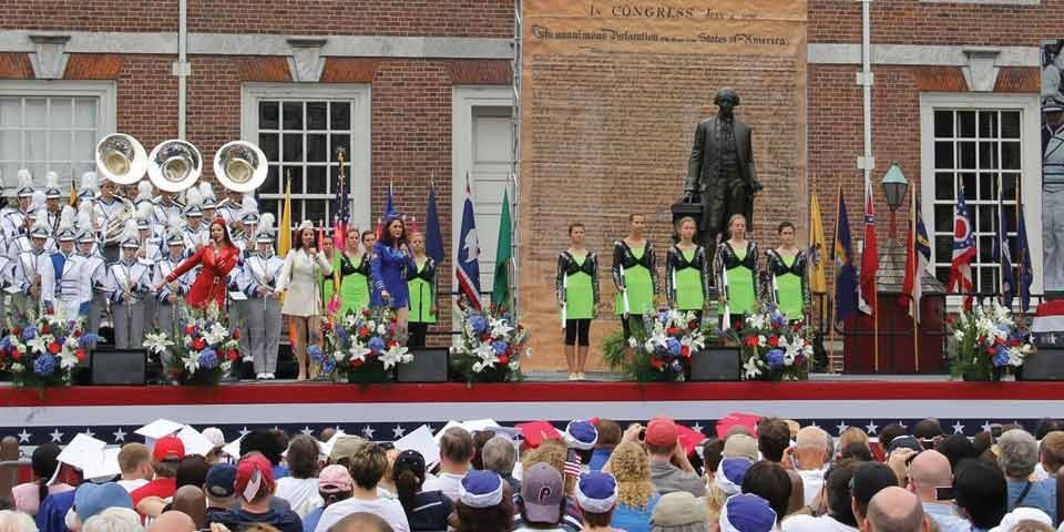 Color photo showing marching band members in silver uniforms on a stage in front of Independence Hall, a red brick building. Color photo showing marching band members in silver uniforms on a stage in front of Independence Hall, a red brick building.