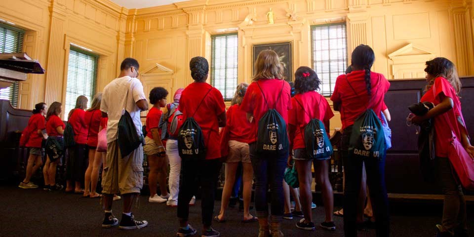 Color photo showing a group of teens in matching red shirts from the back as they look at a room in Independence Hall. Color photo showing a group of teens in matching red shirts from the back as they look at a room in Independence Hall.