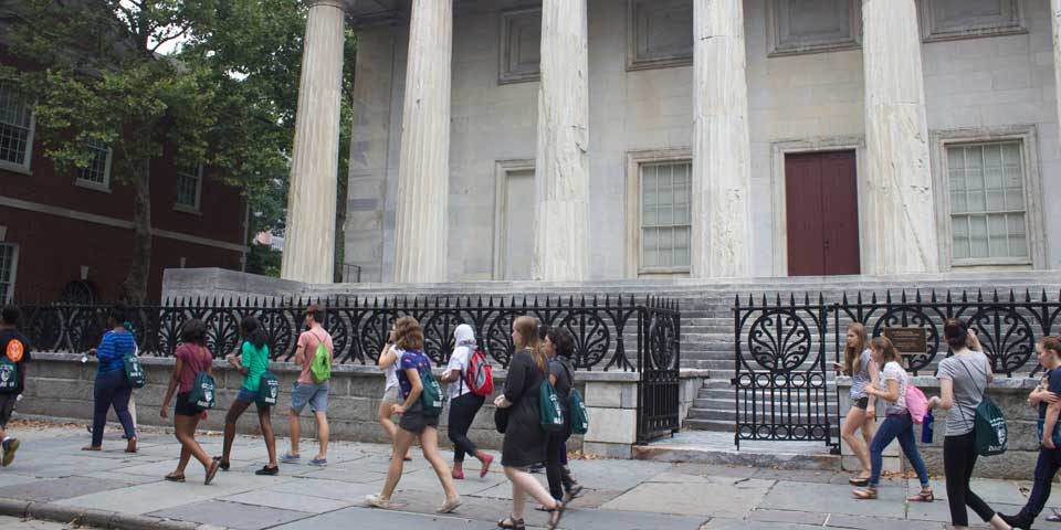 Color photo of a group of teens walking down the sidewalk in front of a large marble-columned building.