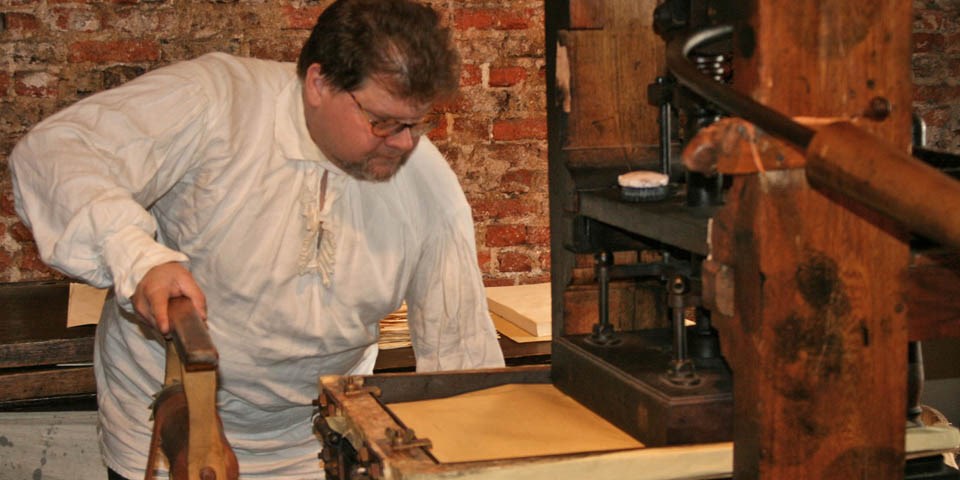 Color photo of man in 18th-century costume demonstrating the printing press.