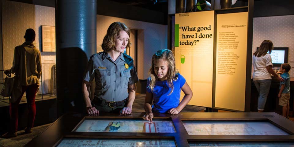 Color photo of female park ranger looking at a screen on an exhibit table with a young girl; exhibit panel with the words "What Good Have I Done Today" is visible behind them.