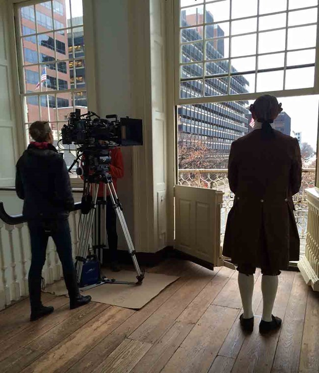 A person with a large camera pointed at a person in 18th Century attire in front of window and open doors to balcony.