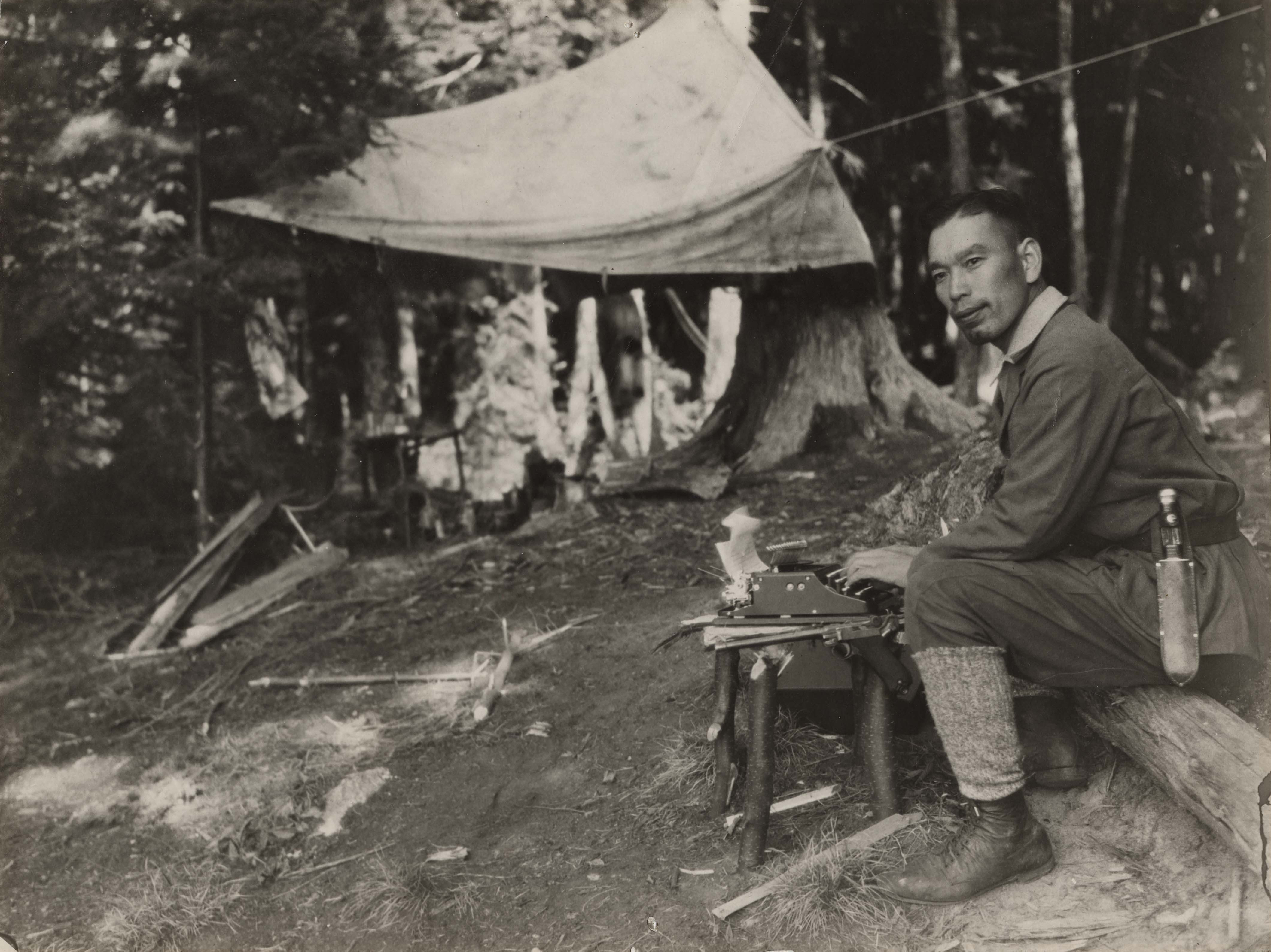 Jun Fujita sits at a camp in a woodland setting. He has a canvas tent, and is seated at a makeshift desk with log legs. A typewriters sits atop the desk. He has a pistol at its side, and is also carrying a large knife on his hip.