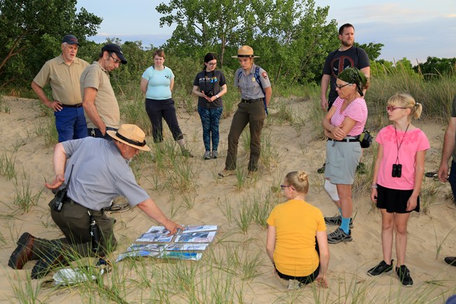 Two national park rangers are surrounded by a group of visitors on the summit of Mount Baldy looking at graphics.