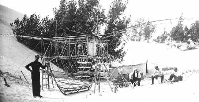 Historic black and white photograph of a camp in sand dunes featuring a canvas tent, 4 men and the wooden frame of an experimental flying machine.