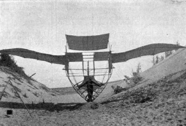Historic black and white photograph of an experimental glider, the Albatross, a large, wooden hulled airship. It rests at the top of a sand dune overlooking Lake Michigan.