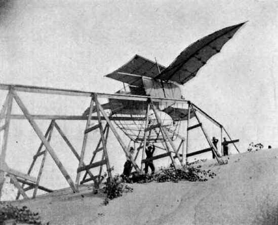 An elevated, wooden track stands at the top of a sand dune with an early experimental aeroplane resting on top with wings outstretched and four individuals reading it. Historic black and white photograph.