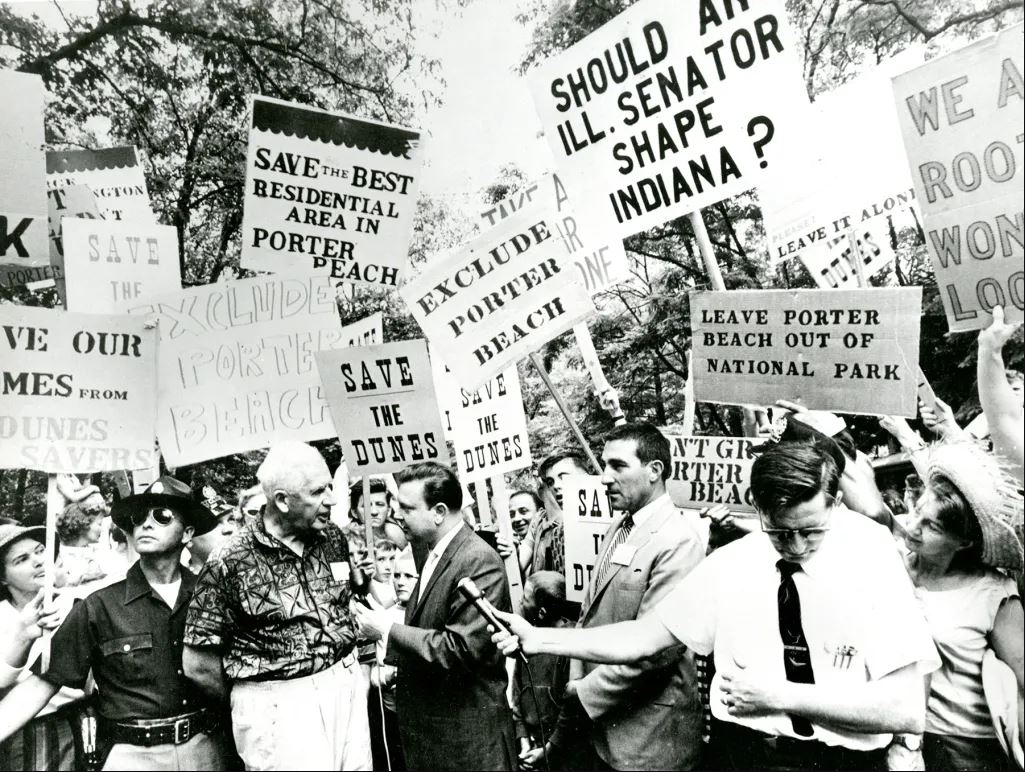 Black and white photograph of Paul Douglas and Secretary Udall surrounded by citizens and press. Citizens holding pro-park and anti-park signage.