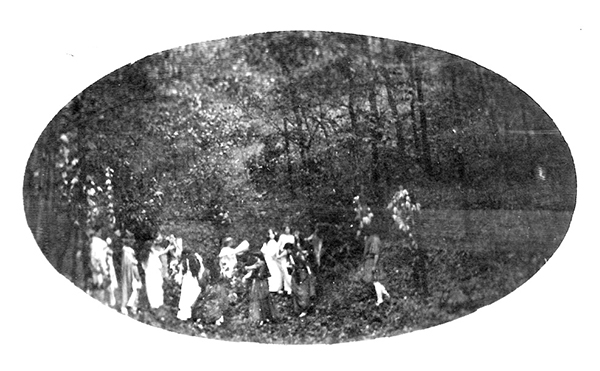 black and white photograph of around a dozen people in mostly light colored outfits amongst a forest scene. They are performing a masque