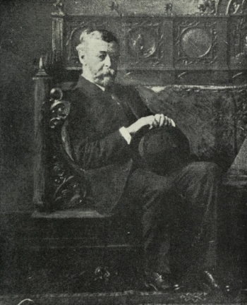 Henry Blake Fuller as an older gentleman Black and white photograph of an elderly gentleman in a dark suit, seated in a dark chair within a dark room.
