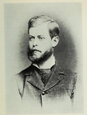 Portrait of a young man in a dark suit with a beard and mustache.