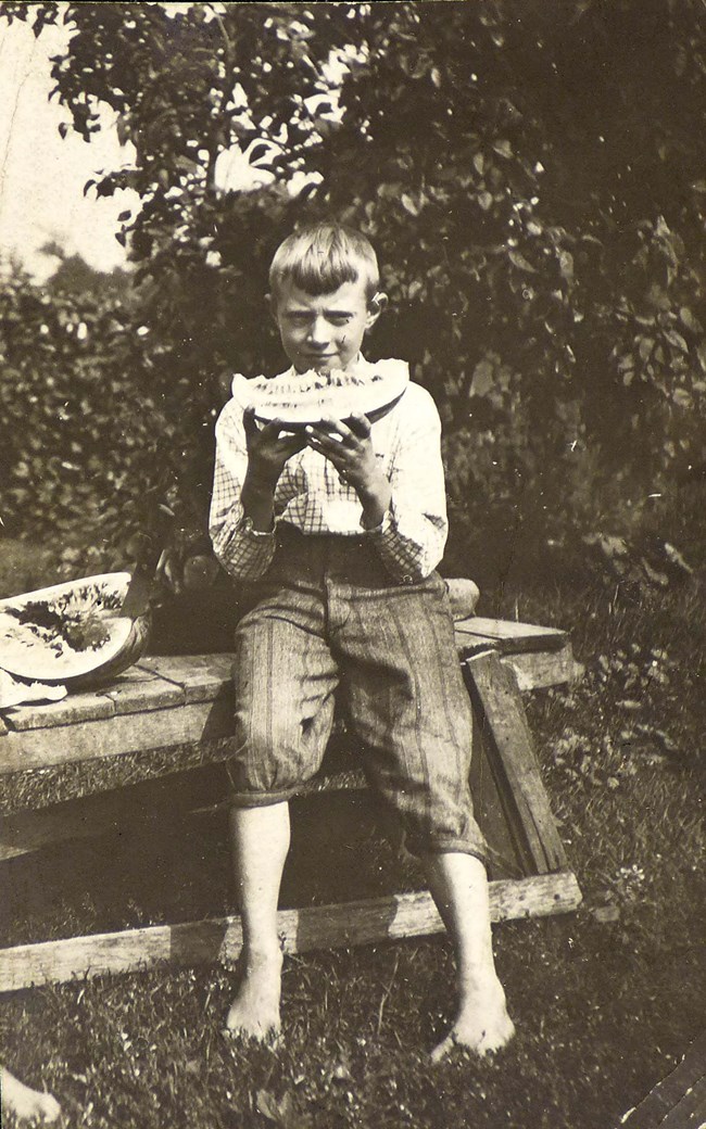 Edwin Way Teale as a boy. A 10-year-old boy sits on a crude wooden table with trousers on and a plaid shirt; eating a melon